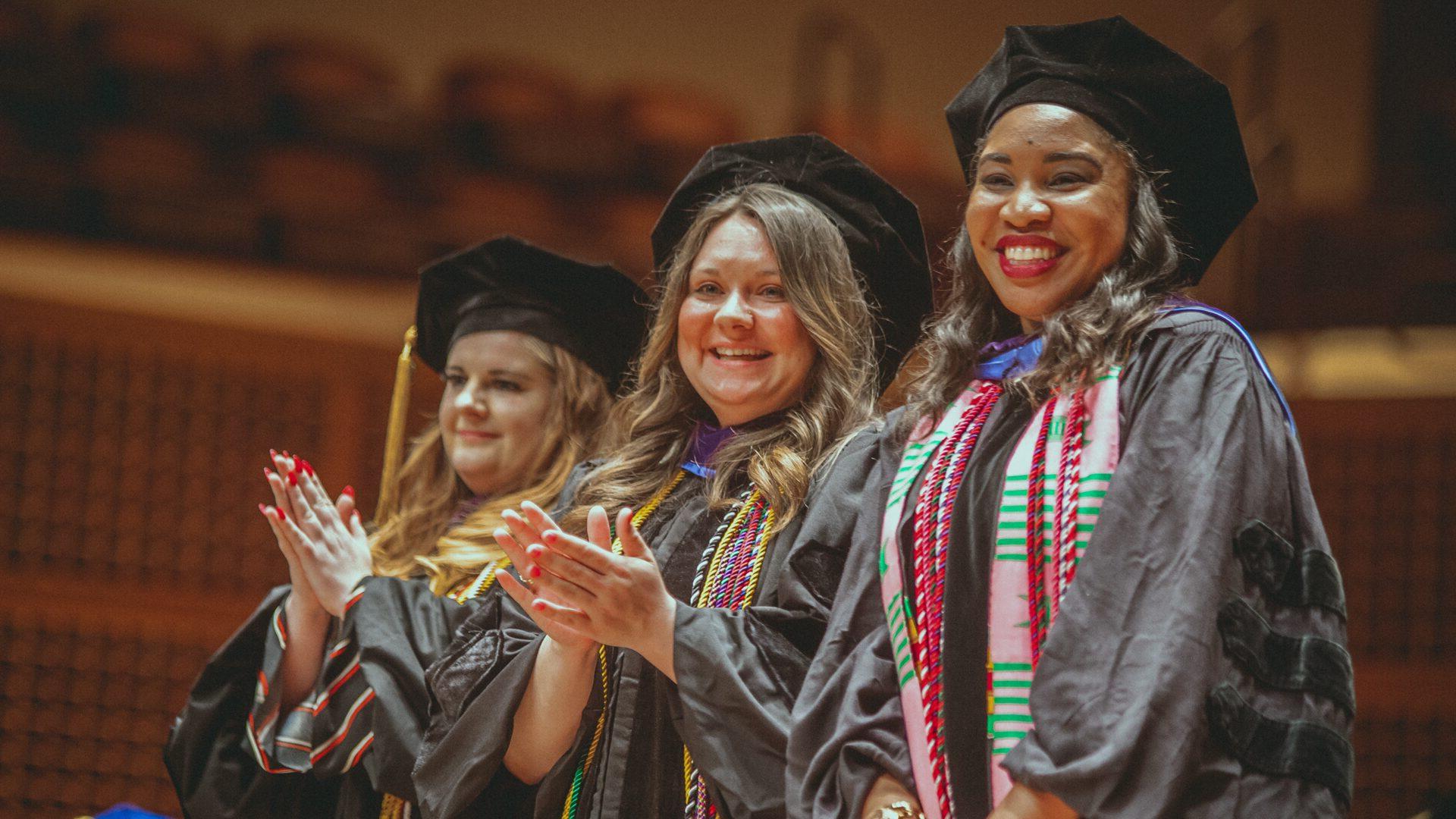 Three graduates in academic regalia smiling and clapping.